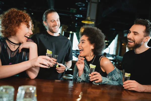 Carefree interracial people holding tequila shots near stand in bar — Stock Photo