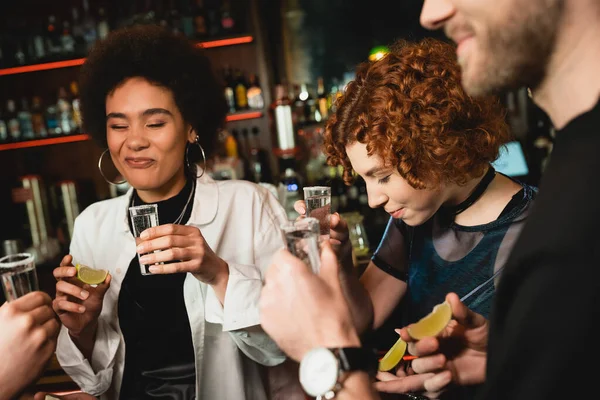 Afro-américaine grimacant après avoir dégusté du citron vert aigre près d'amis multiethniques tenant des tirs de tequila dans un bar — Photo de stock