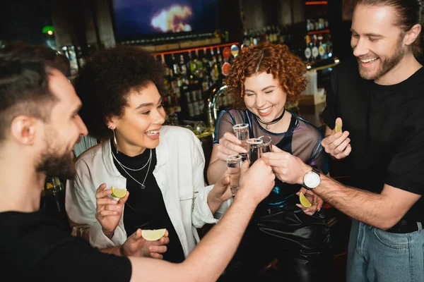 Cheerful multiethnic friends toasting tequila with salt in glasses while holding lime — Stock Photo
