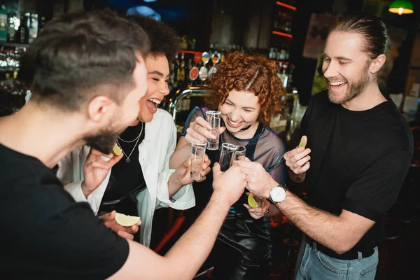 Smiling redhead woman clinking tequila shots with interracial friends in bar — Stock Photo