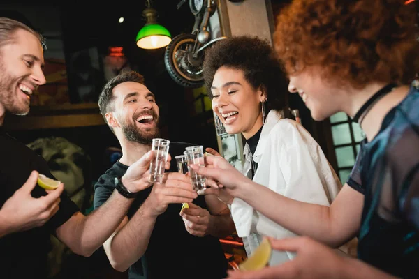 Hombre positivo tintineando tequila con amigos interracial mientras pasa tiempo en el bar - foto de stock