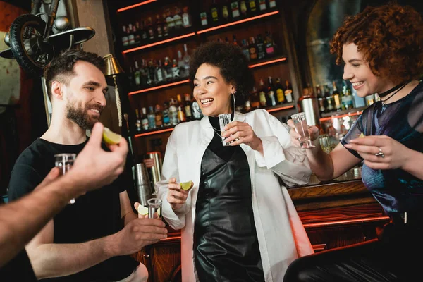 Multiethnic friends holding tequila and fresh lime near stand in bar — Stock Photo