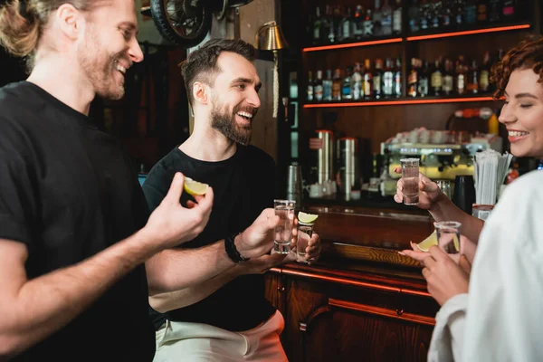 Smiling friends holding tequila shots and ripe lime near stand in bar — Stock Photo