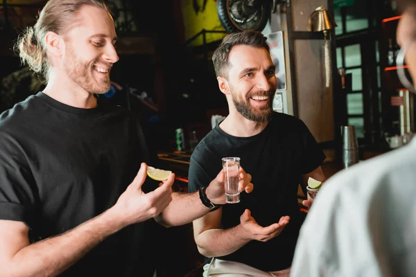 Amigos multiétnicos conversando e segurando tiros de tequila e limão no bar — Fotografia de Stock