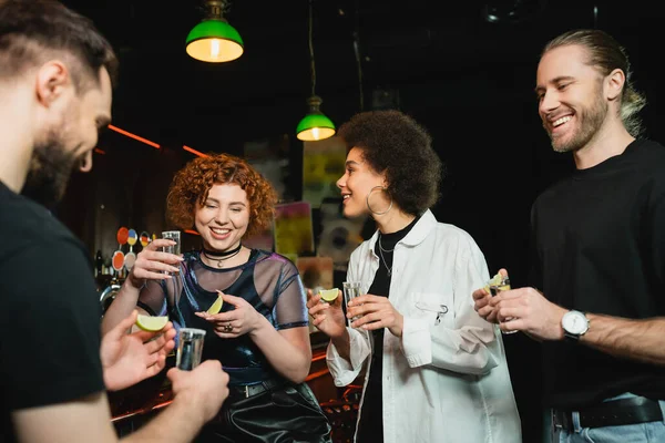 Улыбающиеся молодые межрасовые женщины, держащие текилу и лайм рядом с друзьями в баре ночью — стоковое фото