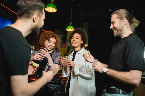 Pessoas multiétnicas positivas conversando e segurando tequila e limão fresco no bar — Fotografia de Stock