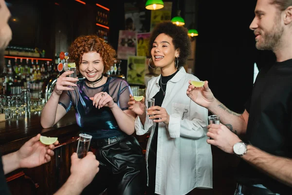 Cheerful multiethnic people having fun and holding tequila shots in bar — Stock Photo
