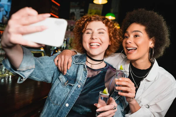 Mulher positiva tomando selfie com amigo afro-americano e segurando tequila no bar — Fotografia de Stock