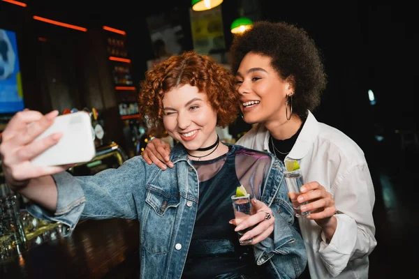 Carefree multiethnic girlfriends taking selfie and holding tequila in bar — Stock Photo
