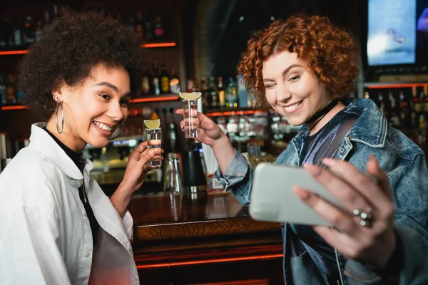 Novia multiétnica positiva sosteniendo tequila con lima y tomando selfie en el bar - foto de stock
