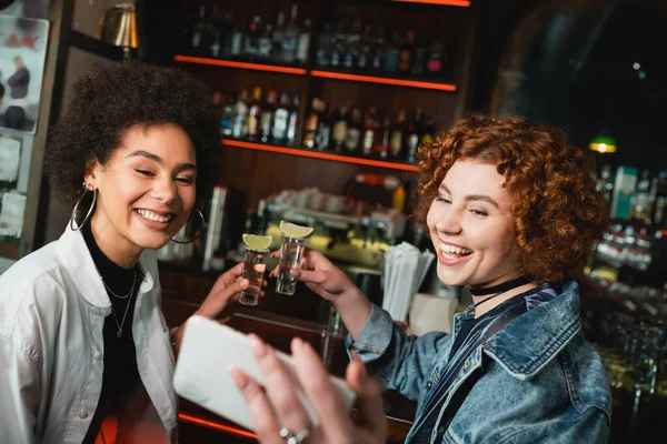 Smiling multiethnic friends holding tequila with lime and taking selfie in bar at night — Stock Photo