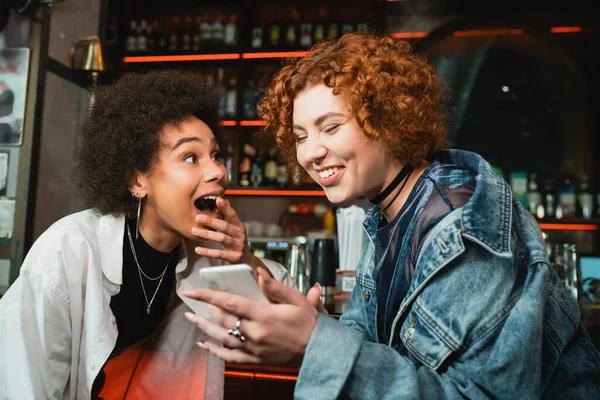 Femme rousse excitée utilisant un smartphone près d'un ami afro-américain dans un bar — Photo de stock