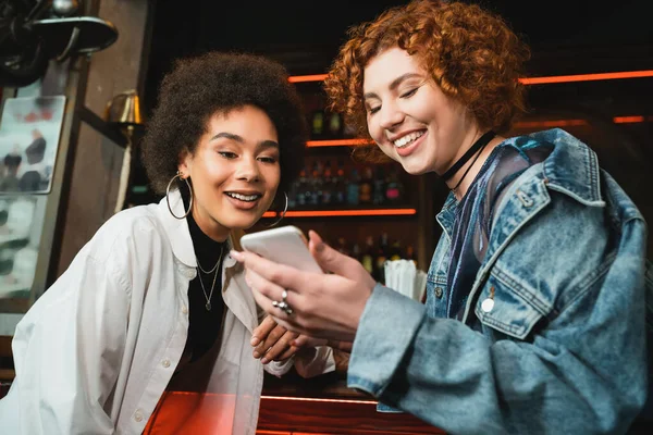 Amigos multiétnicos positivos utilizando el teléfono inteligente cerca de stand en el bar por la noche - foto de stock