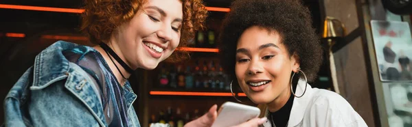 Positivo interracial de las mujeres jóvenes utilizando el teléfono celular en el bar, pancarta - foto de stock