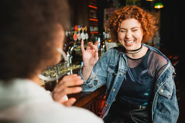 Mulher ruiva positiva sentado perto de amigo afro-americano borrado e ficar no bar — Fotografia de Stock