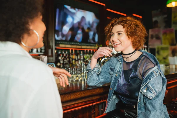 Junge rothaarige Frau spricht mit verschwommenem afrikanisch-amerikanischem Freund in der Nähe eines Stands in einer Bar — Stockfoto