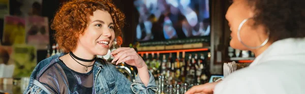 Cheerful redhead woman talking to blurred african american friend in bar, banner — Stock Photo