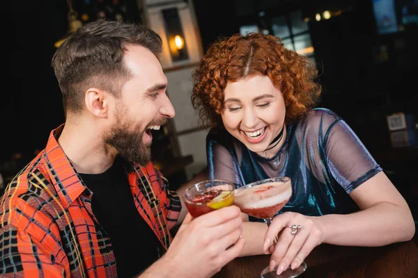 Mujer pelirroja alegre riendo y sosteniendo cóctel de espuma cerca de amigo barbudo en el bar - foto de stock