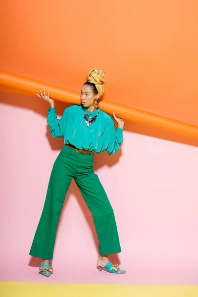 Mujer afroamericana de moda en pañuelo para la cabeza y traje brillante posando sobre fondo colorido - foto de stock