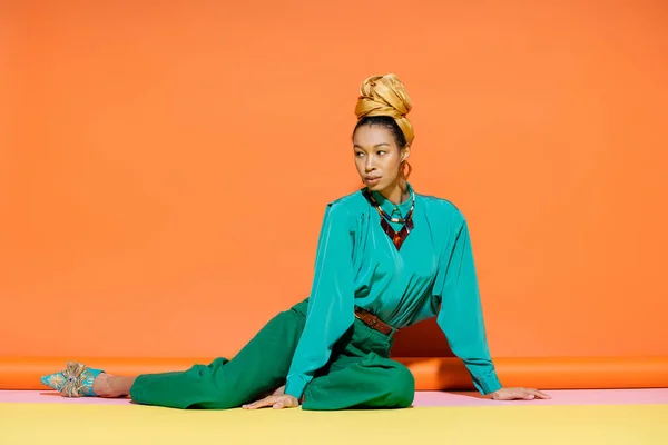 Stylish african american woman in headscarf and bright blouse sitting on colorful background — Stock Photo