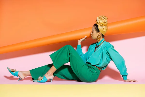 Vista lateral del modelo afroamericano de moda en traje de verano sentado sobre un fondo colorido - foto de stock