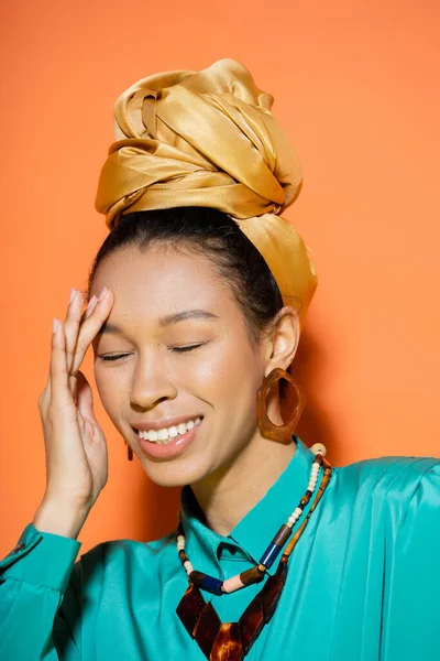 Portrait of smiling african american woman in golden headband touching face on orange background — Stock Photo