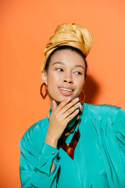 Smiling african american woman in golden headscarf touching chin on orange background — Stock Photo
