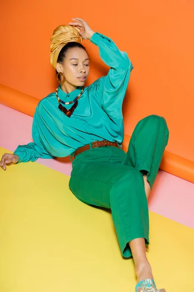 Trendy modello afroamericano in velo e vestiti estivi seduto su sfondo colorato — Foto stock