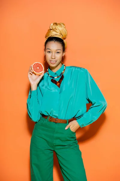 Sonriente modelo afroamericano en traje de verano con pomelo sobre fondo naranja - foto de stock