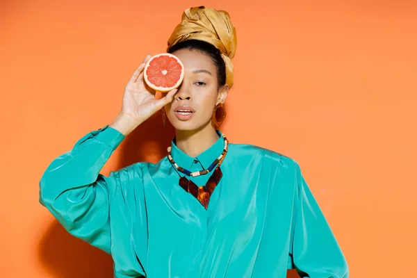 Mujer afroamericana de moda con pañuelo para la cabeza sosteniendo pomelo sobre fondo naranja - foto de stock