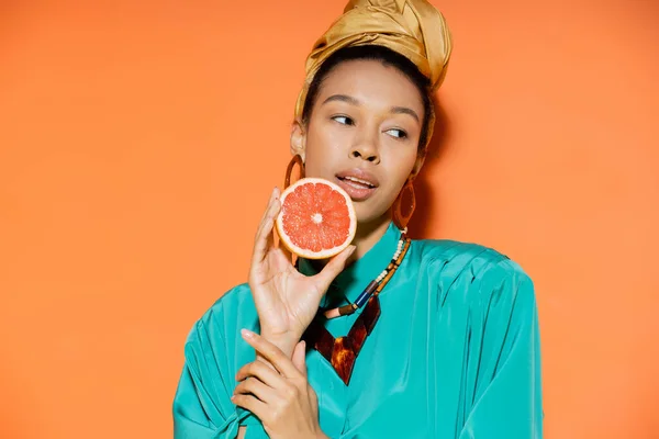 Joven y elegante modelo afroamericano sosteniendo pomelo maduro sobre fondo naranja - foto de stock