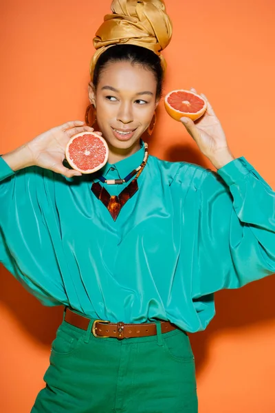 Overjoyed african american woman in summer clothes holding cut grapefruit on orange background — Stock Photo