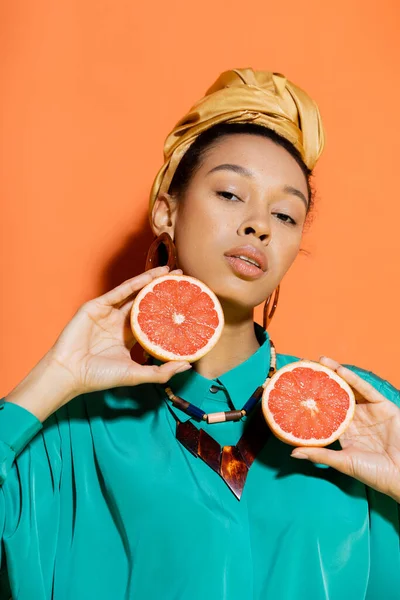 Retrato de elegante modelo afroamericano con pañuelo en la cabeza con toronja cortada sobre fondo naranja - foto de stock