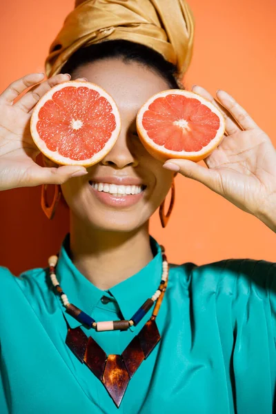 Positive and stylish african american model covering face with grapefruit on orange background — Stock Photo