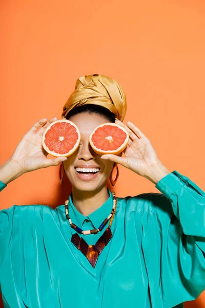 Mujer afroamericana positiva en traje de verano cubriendo la cara con pomelo fresco sobre fondo naranja - foto de stock