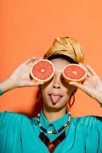 Stylish african american woman sticking out tongue and holding cut grapefruit on orange background — Stock Photo