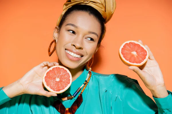 Portrait de femme afro-américaine positive en chemisier tenant du pamplemousse coupé sur fond orange — Photo de stock