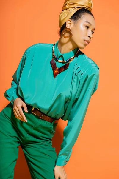 Modelo afroamericano de moda en pañuelo para la cabeza y blusa mirando hacia otro lado sobre fondo naranja - foto de stock
