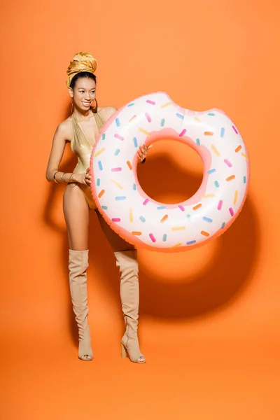Fröhliche und stilvolle afrikanisch-amerikanische Frau im Badeanzug mit Pool-Ring auf orangefarbenem Hintergrund — Stockfoto