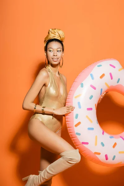 Smiling african american woman in golden swimsuit holding pool ring on orange background — Stock Photo