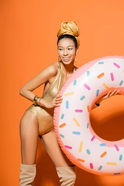 Unbekümmertes afrikanisch-amerikanisches Model im goldenen Badeanzug mit Pool-Ring auf orangefarbenem Hintergrund — Stockfoto