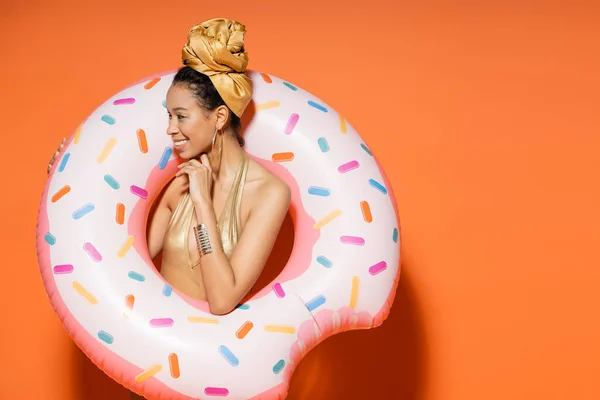 Sonriente mujer afroamericana en pañuelo dorado sosteniendo el anillo de la piscina sobre fondo naranja - foto de stock