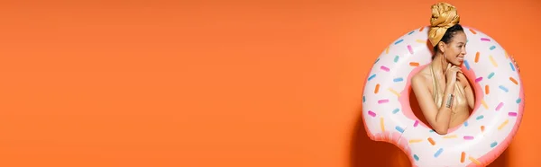 Joyeux modèle afro-américain en foulard doré posant avec anneau de piscine sur fond orange, bannière — Photo de stock