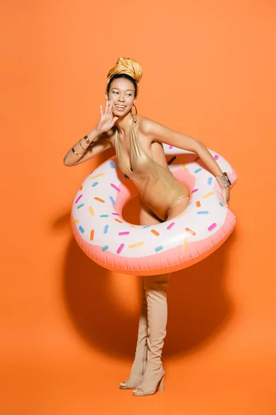 Positives afrikanisch-amerikanisches Model in Kniestiefeln und Badeanzug mit Pool-Ring auf orangefarbenem Hintergrund — Stockfoto