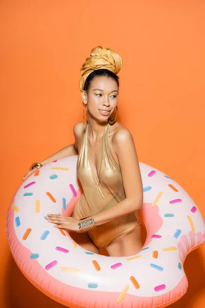 Modèle afro-américain souriant en maillot de bain doré et foulard tenant une bague de piscine sur fond orange — Photo de stock