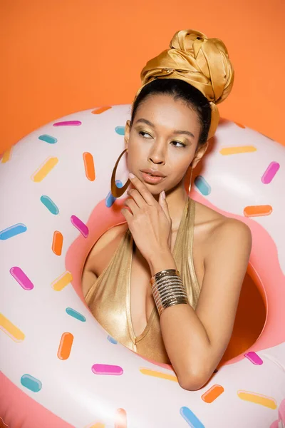 Retrato de mujer afroamericana pensativa en pañuelo para la cabeza posando con anillo de piscina sobre fondo naranja - foto de stock