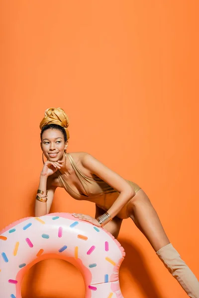 Sonriente mujer afroamericana en traje de baño posando cerca del anillo de la piscina sobre fondo naranja - foto de stock