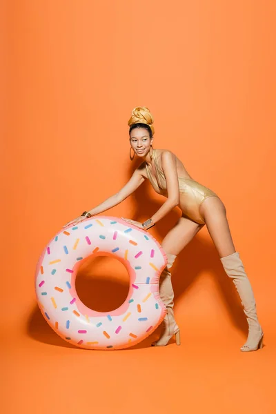 Modelo afroamericano sonriente en botas de rodilla y traje de baño posando cerca del anillo de la piscina sobre fondo naranja - foto de stock