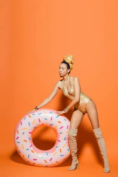 Full length of stylish african american model in golden swimsuit standing near pool ring on orange background — Stock Photo