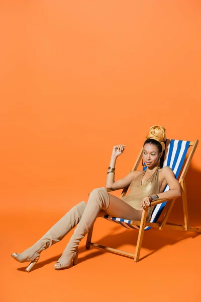 Full length of stylish african american woman in swimsuit and headscarf sitting on deck chair on orange background — Stock Photo